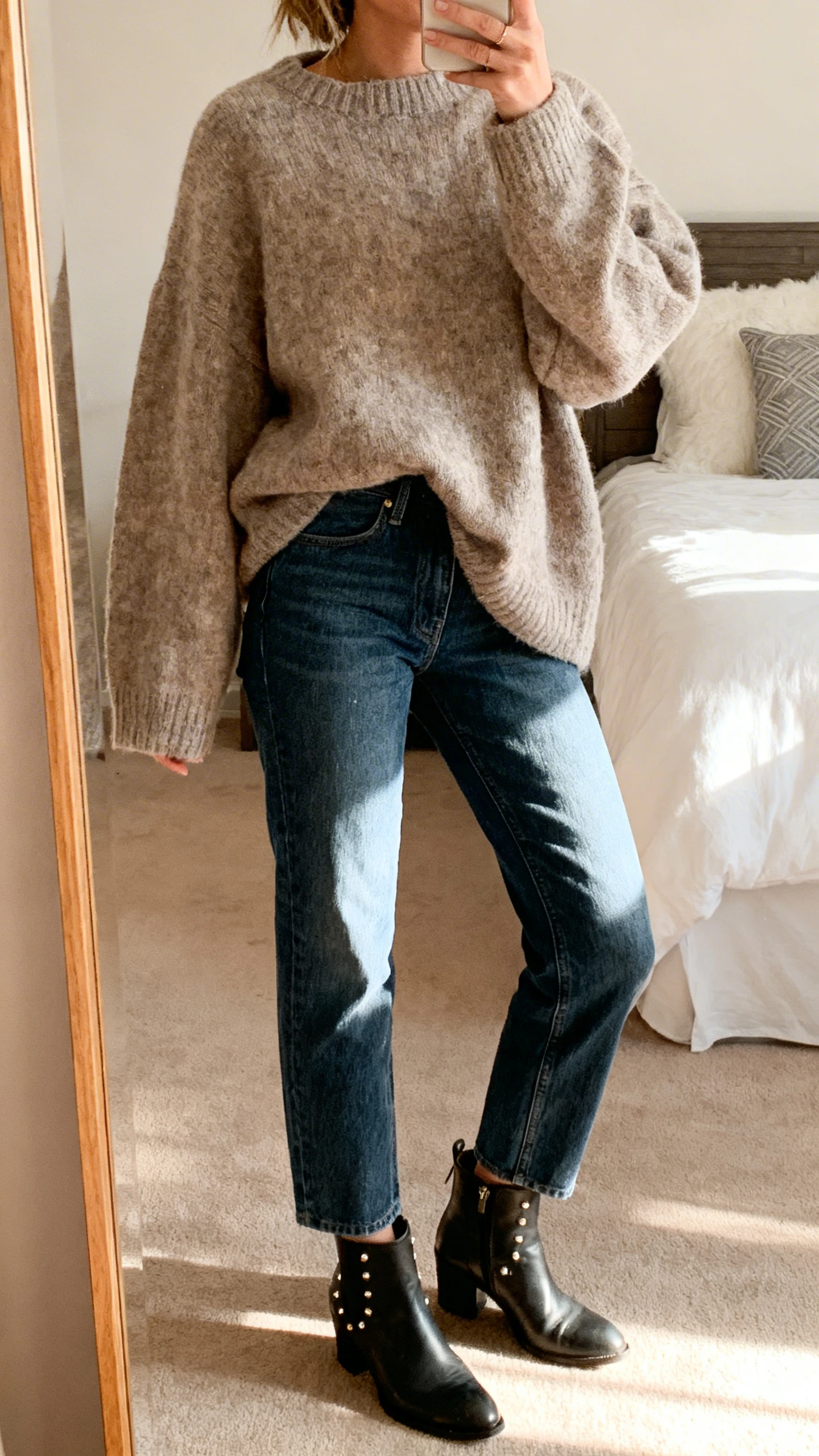 iPhone mirror selfie of a woman wearing an oversized sweater with straight-leg jeans and ankle boots, minimal studs, face not visible, bedroom mirror, cozy afternoon light, iPhone photo quality.