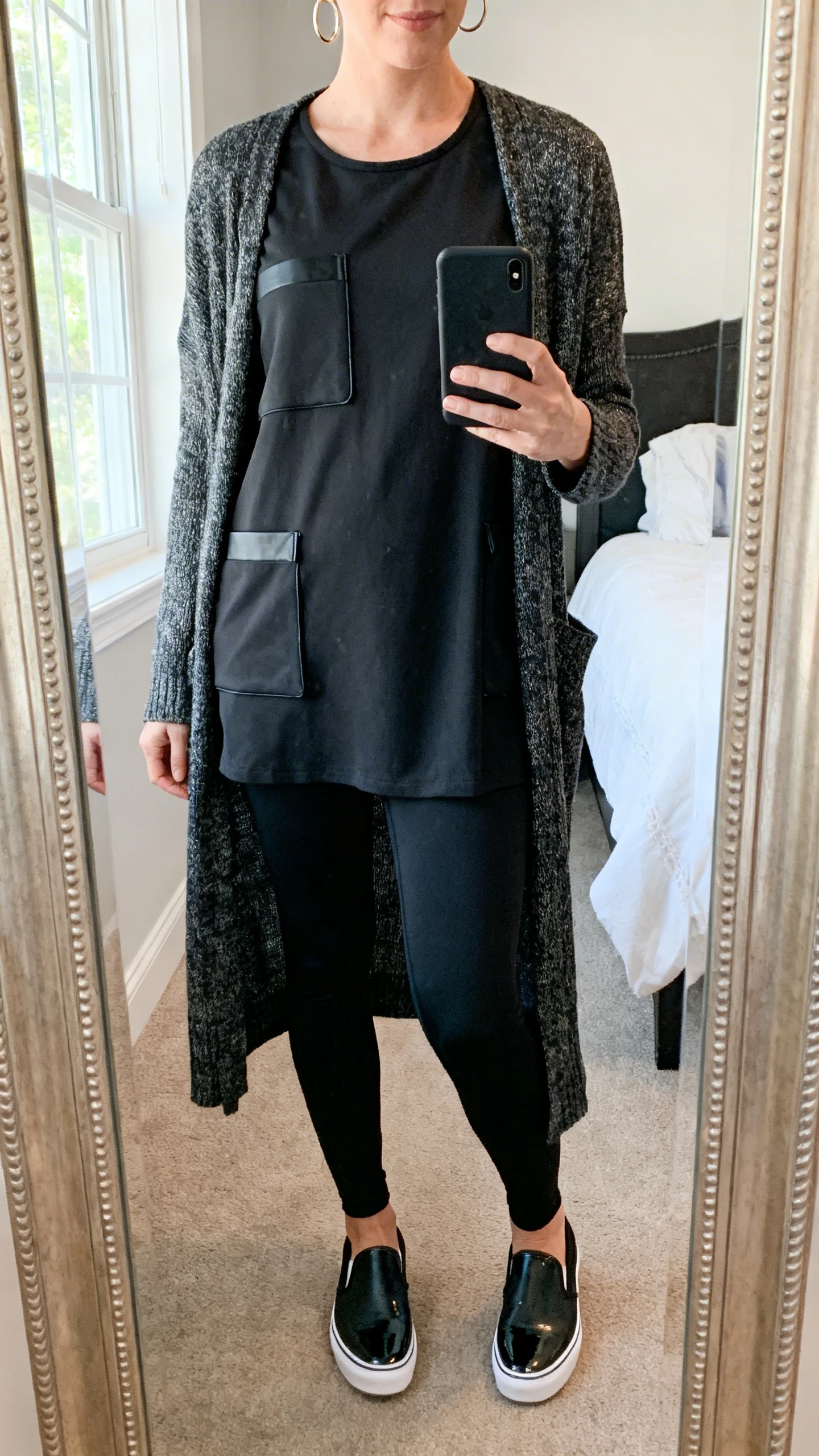 iPhone mirror selfie of a woman wearing black leggings and a structured tunic-length top with pockets, layered with a longline cardigan, sporty-luxe slip-on shoes, and small hoop earrings, face not visible, cropped at shoulders, bedroom mirror, natural window light, iPhone photo quality.