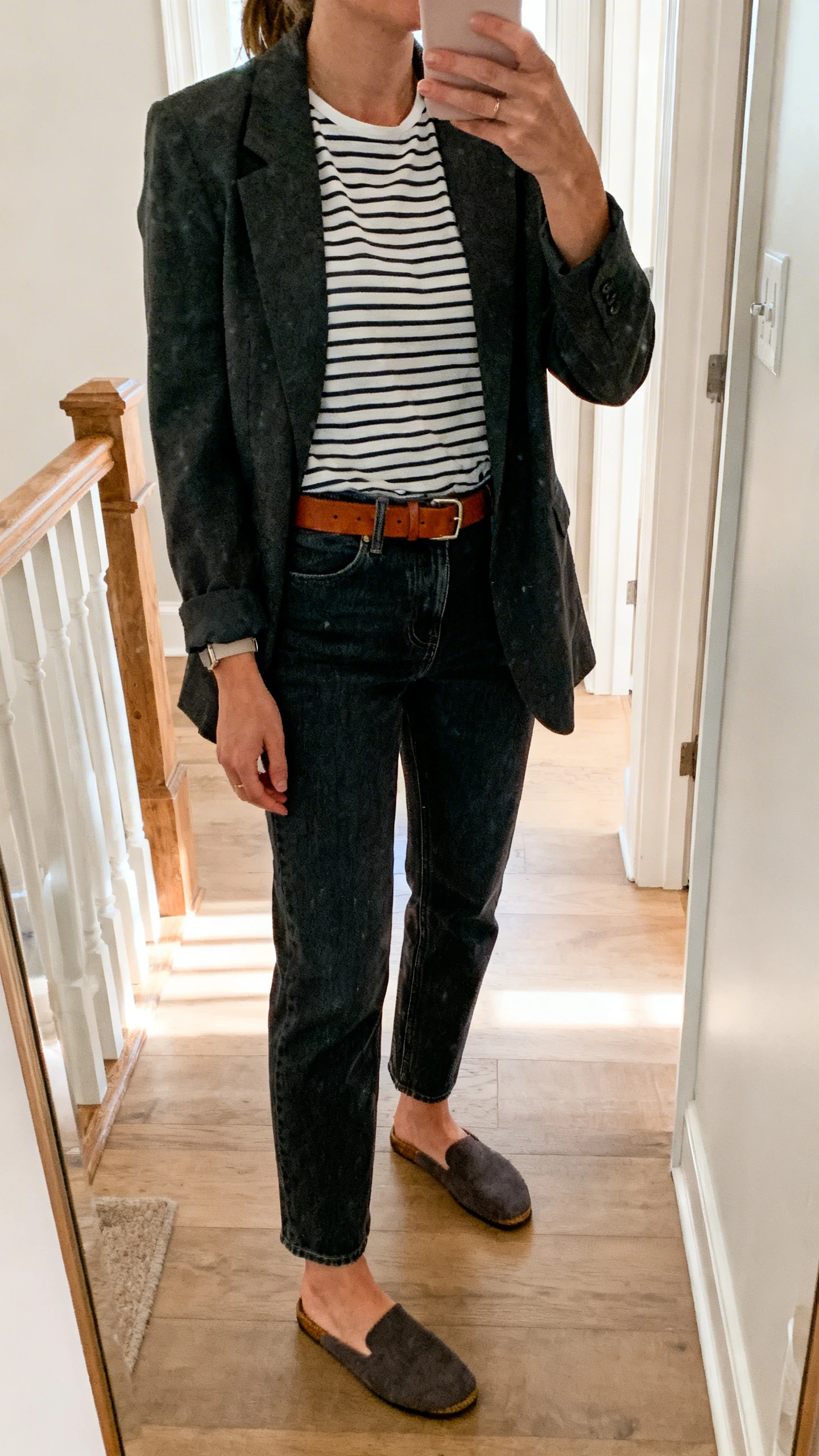 iPhone mirror selfie of a woman wearing dark-wash straight-leg jeans with a tucked-in striped tee, relaxed blazer, slip-on mules, and a leather belt, face not visible, cropped at neck, entryway mirror, morning light, iPhone photo quality.