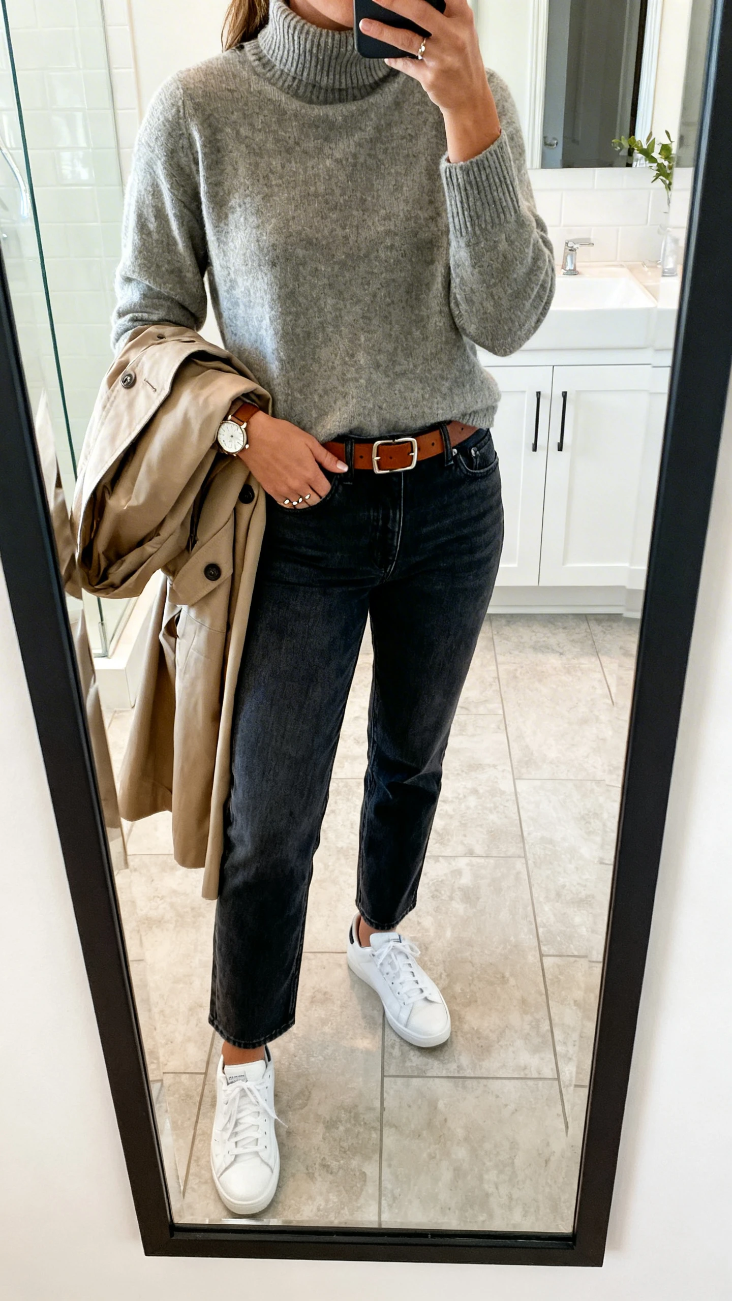 iPhone mirror selfie of a woman wearing dark-wash straight-leg jeans with a lightweight mock-neck sweater, white sneakers, a leather belt, simple studs, a watch, and a trench coat draped over one arm, face not visible, bathroom mirror, natural indoor light, iPhone photo quality.