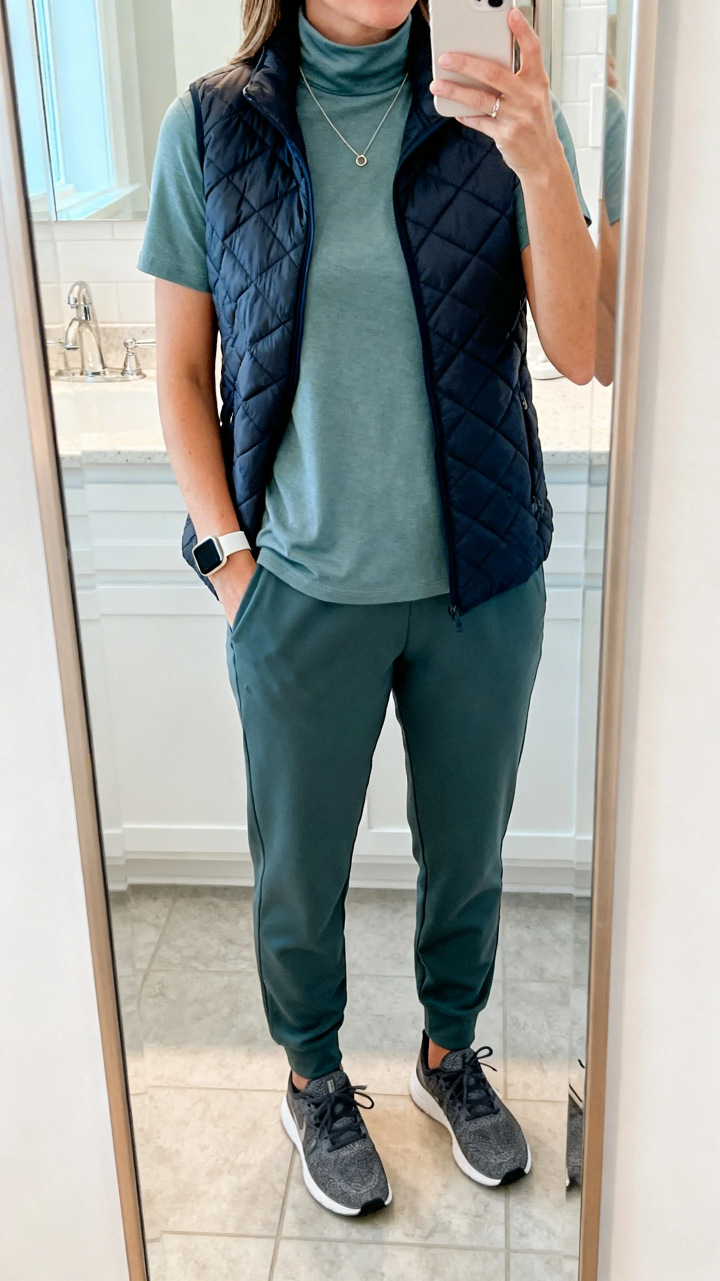 iPhone mirror selfie of a woman wearing tailored joggers, a smooth mock-neck tee, a quilted vest, low-profile trainers, and a minimalist necklace with a smartwatch, face not visible, cropped at neck, bathroom mirror, bright indoor lighting, iPhone photo quality.