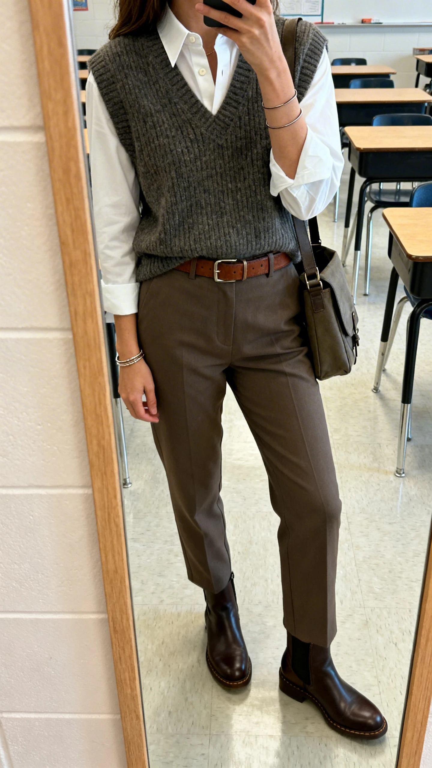 iPhone side-angle mirror selfie of a woman in a knit sweater vest (solid) over a white tailored button-down, straight-leg trousers, Chelsea boots, leather belt, thin bangles, and a messenger bag, face not visible, cropped at shoulders, classroom mirror, natural indoor light, iPhone photo quality.