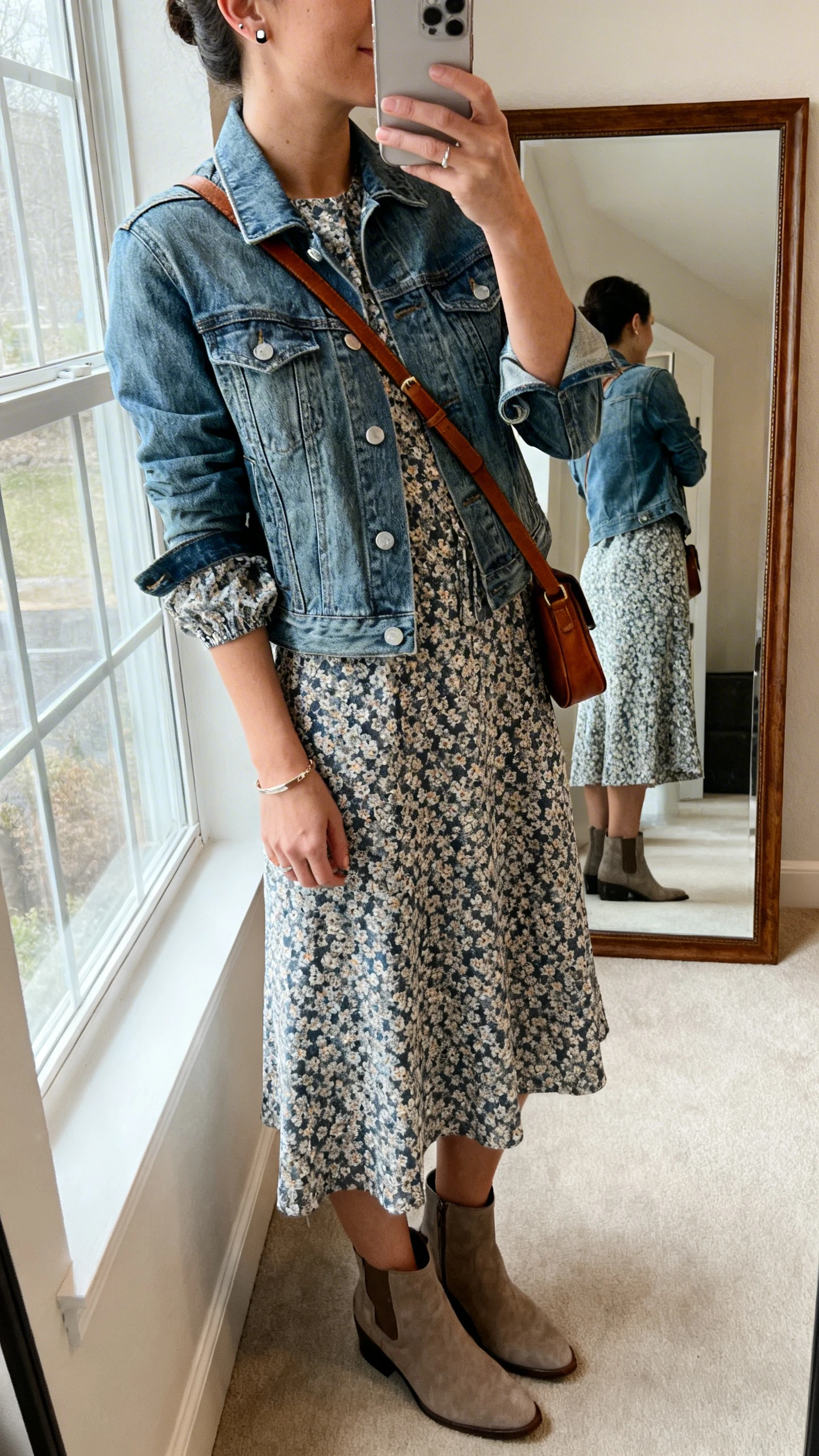 iPhone side-angle mirror selfie of a woman in a printed midi dress with sleeves, medium-wash denim jacket, ankle boots, a leather crossbody, subtle bracelet, and stud earrings, face not visible, full-length mirror near a window, soft daylight, iPhone photo quality.