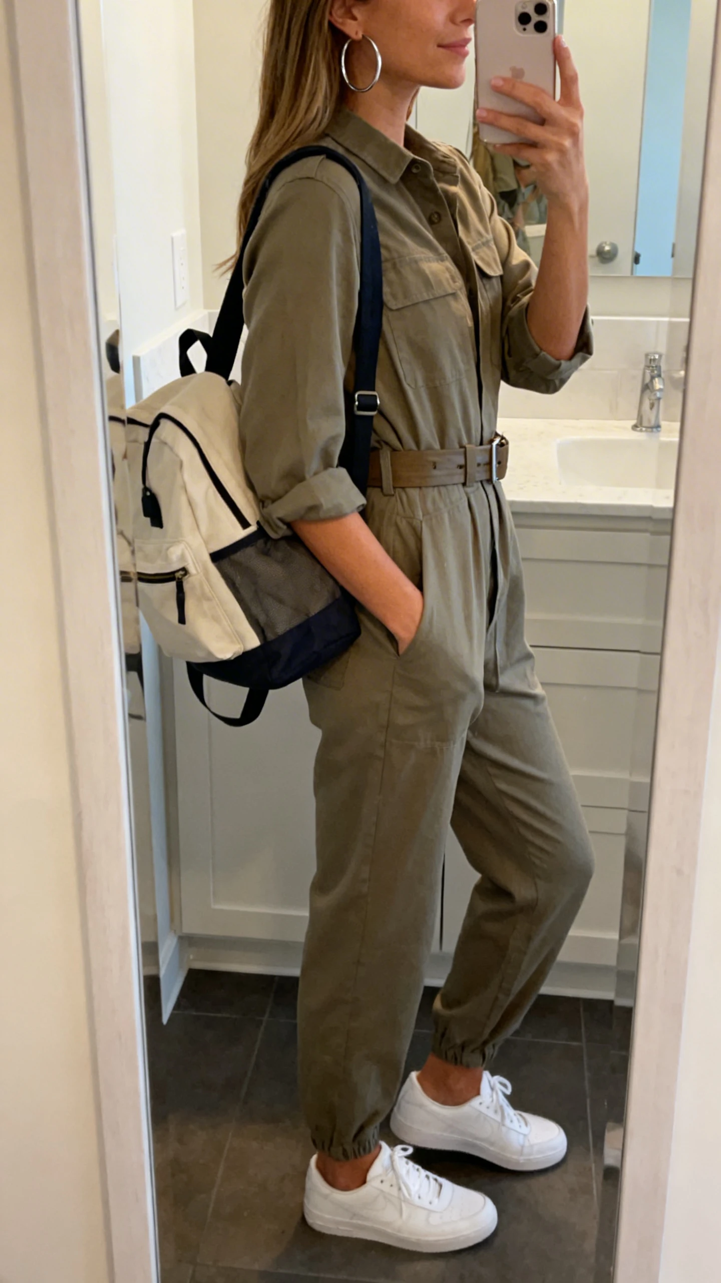 iPhone side-angle mirror selfie of a woman in a utility-style jumpsuit cinched with a belt, white sneakers, hoop earrings, and a backpack tote, face not visible, bathroom mirror, soft indoor lighting, iPhone photo quality.