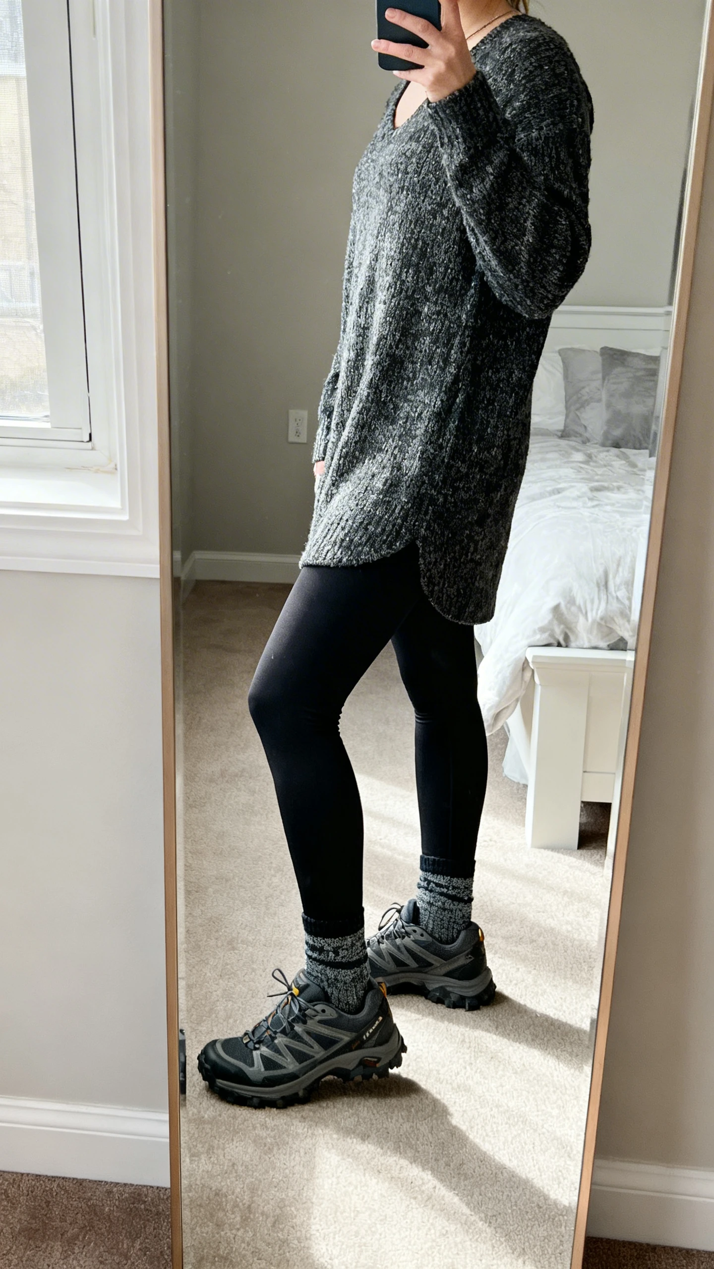 iPhone side-angle mirror selfie of a woman in black leggings, a cozy tunic sweater, all-terrain sneakers, and sporty ankle socks, face not visible, cropped at neck, bedroom mirror, natural window light, iPhone photo quality.