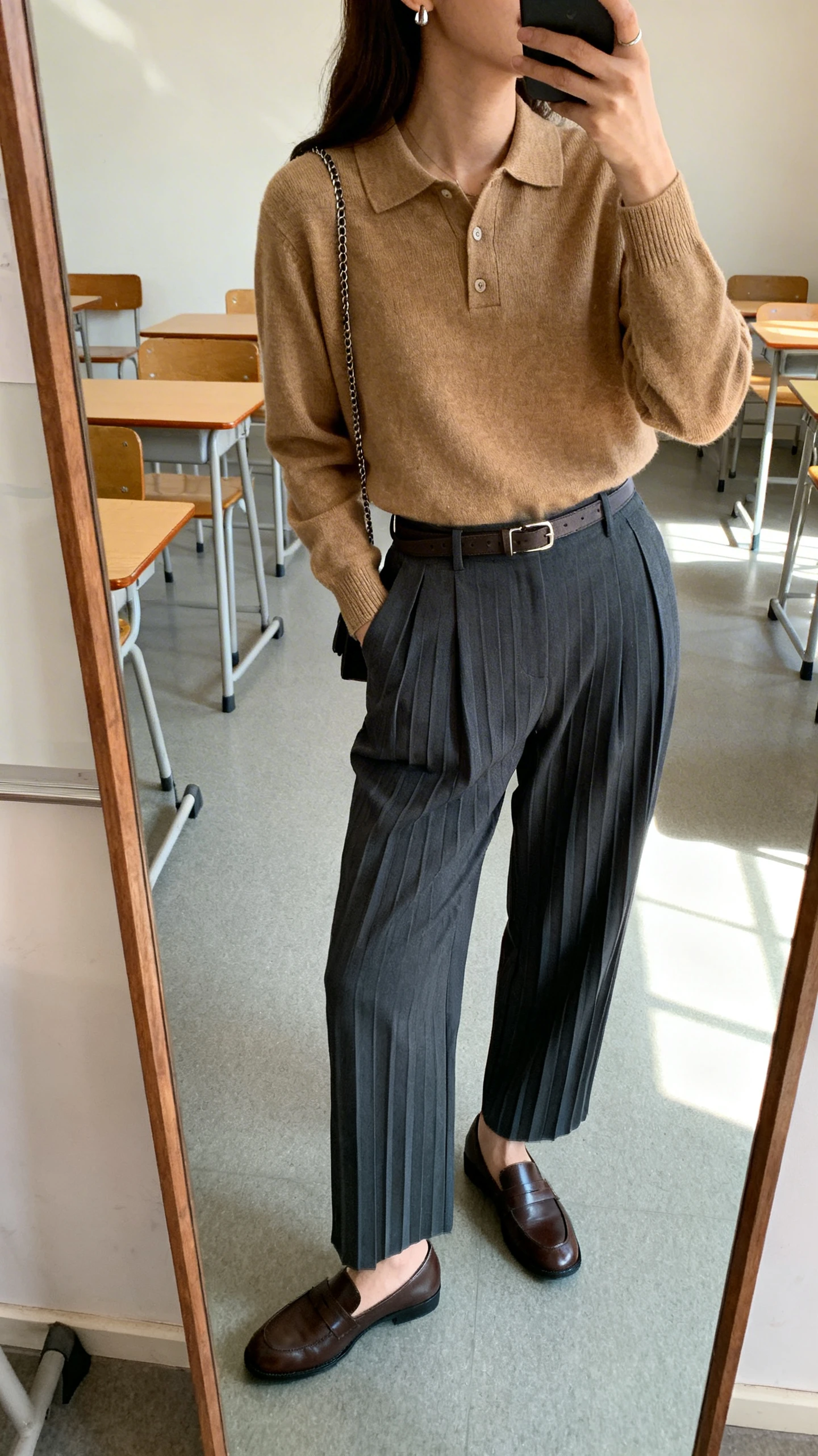 iPhone side-angle mirror selfie of a woman in high-waisted charcoal pleated trousers with a tan knit polo, leather loafers, a minimal belt, subtle chain, stud earrings, and a neat crossbody, face not visible, cropped at neck, classroom-like corner with full-length mirror, soft morning light, iPhone photo quality.