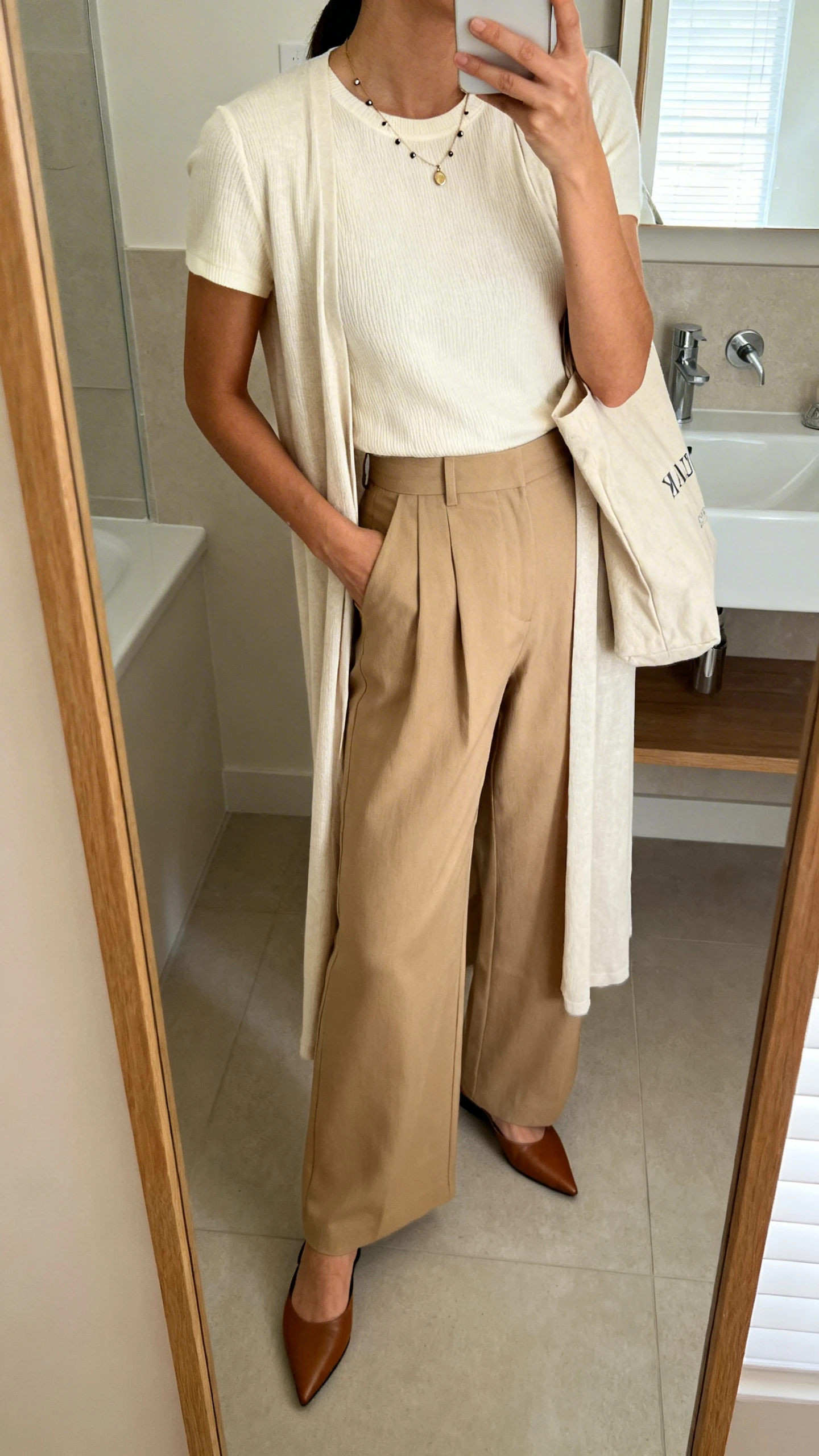iPhone side-angle mirror selfie of a woman wearing high-waisted wide-leg trousers in sand, a cream short-sleeve knit tee, a lightweight longline cardigan, pointed loafer mules, a minimalist pendant necklace with studs, and a canvas tote, face not visible, bathroom mirror, indoor natural light, iPhone photo quality.