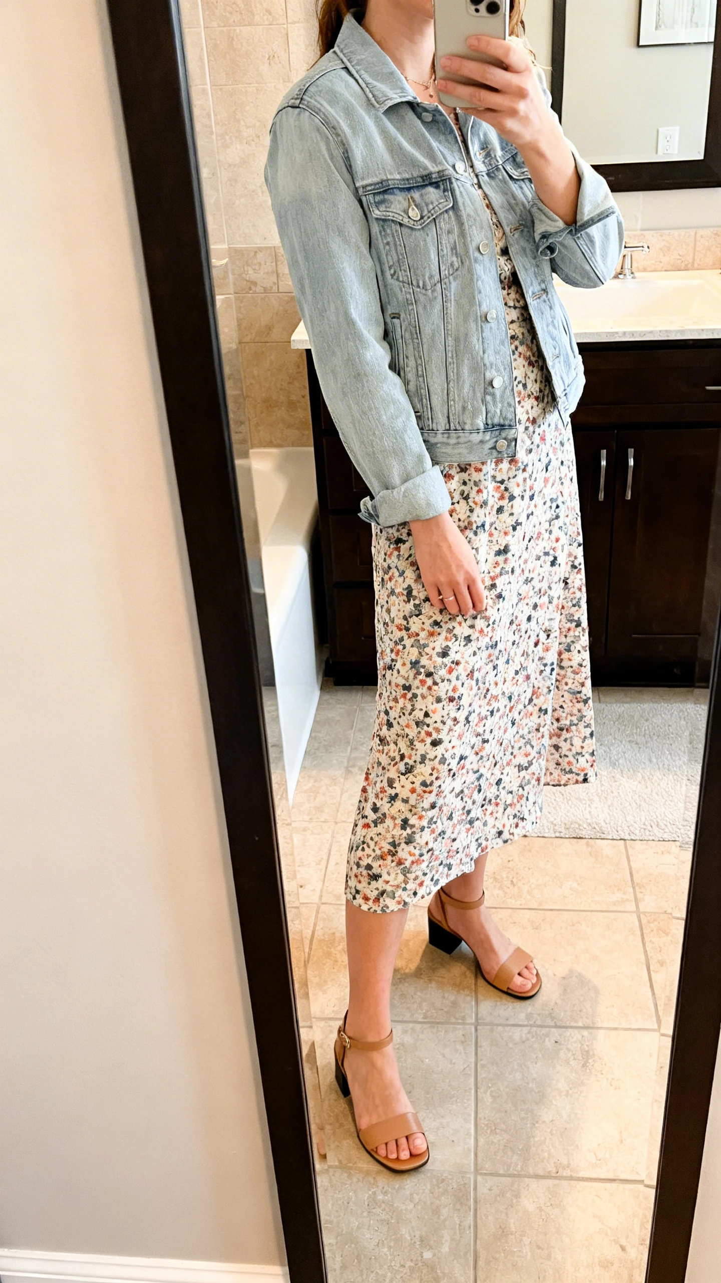 iPhone side-angle mirror selfie of a woman wearing a floral midi dress with a light-wash denim jacket and low block-heel sandals, face not visible, bathroom full-length mirror, morning indoor lighting, iPhone photo quality.