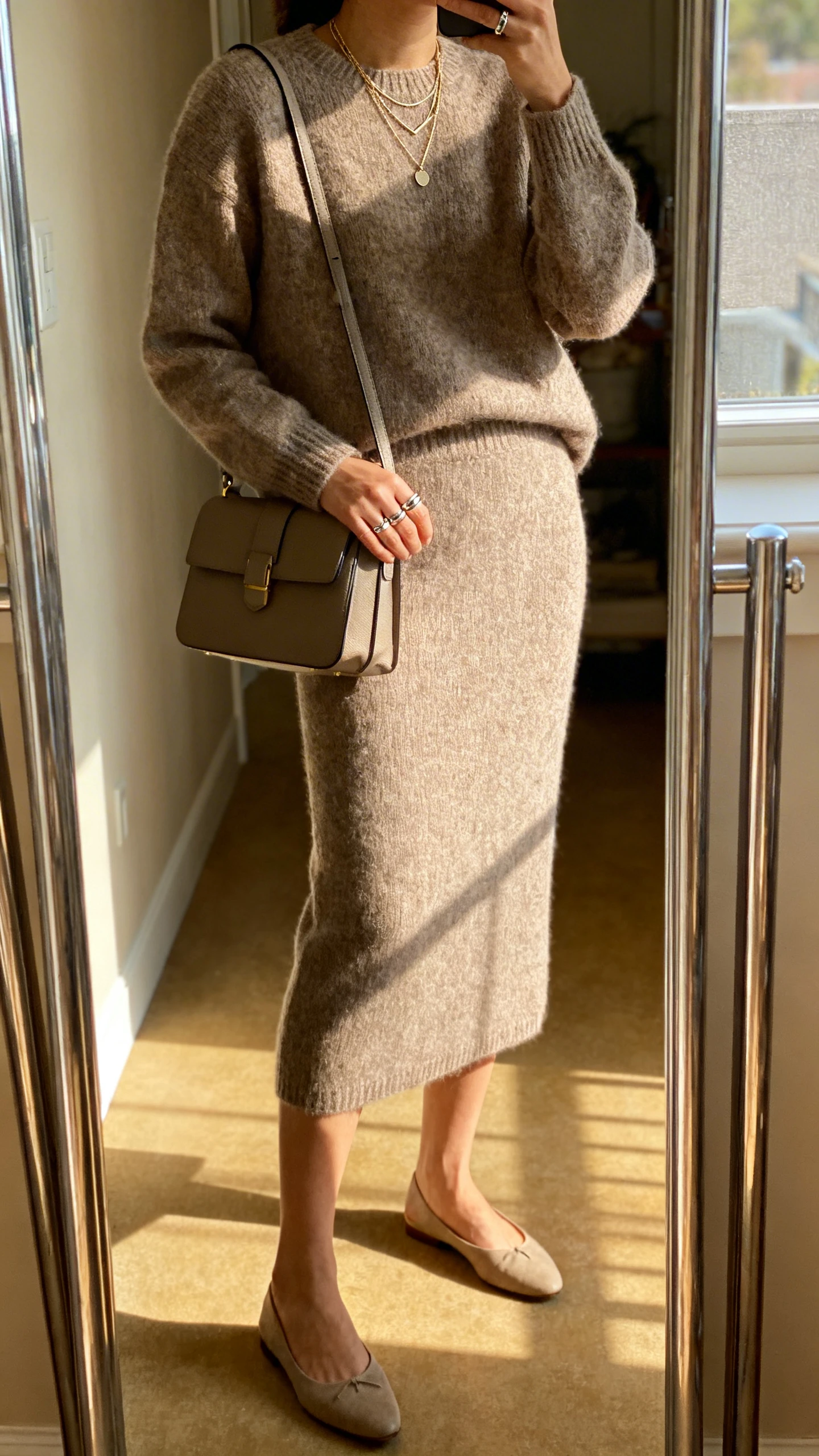iPhone side-angle mirror selfie of a woman wearing a coordinated knit sweater and midi skirt set, almond-toe flats, delicate layered necklaces, simple stacked rings, and a structured satchel, face not visible, cropped at shoulders, full-length mirror in hallway, soft afternoon window light, iPhone photo quality.
