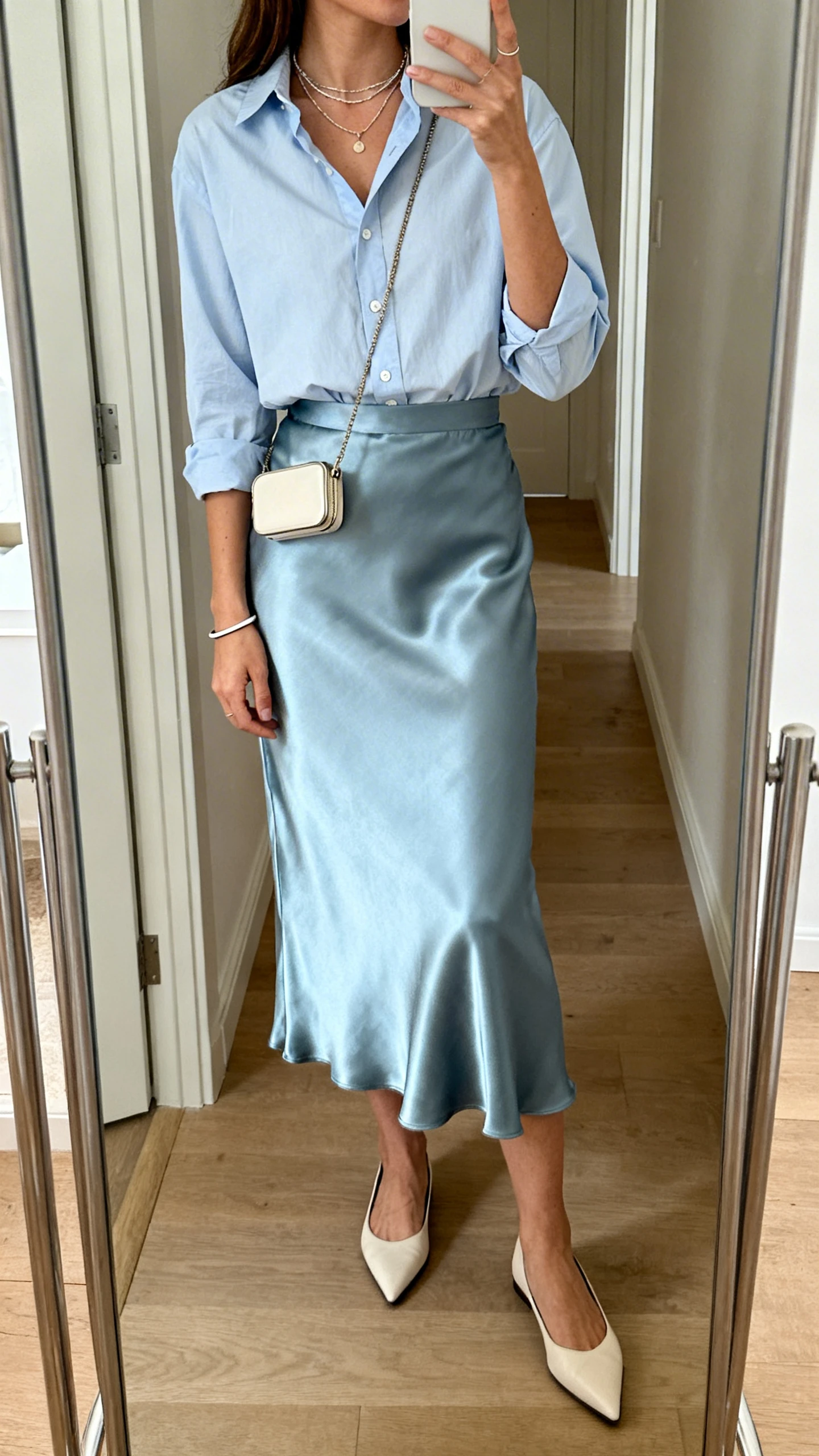 iPhone side-angle mirror selfie of a woman wearing a light blue button-down shirt tucked into a satin midi skirt, pointed-toe flats, delicate layered necklaces, a thin bracelet, and a compact crossbody, face not visible, full-length mirror in hallway, soft daylight, iPhone photo quality.
