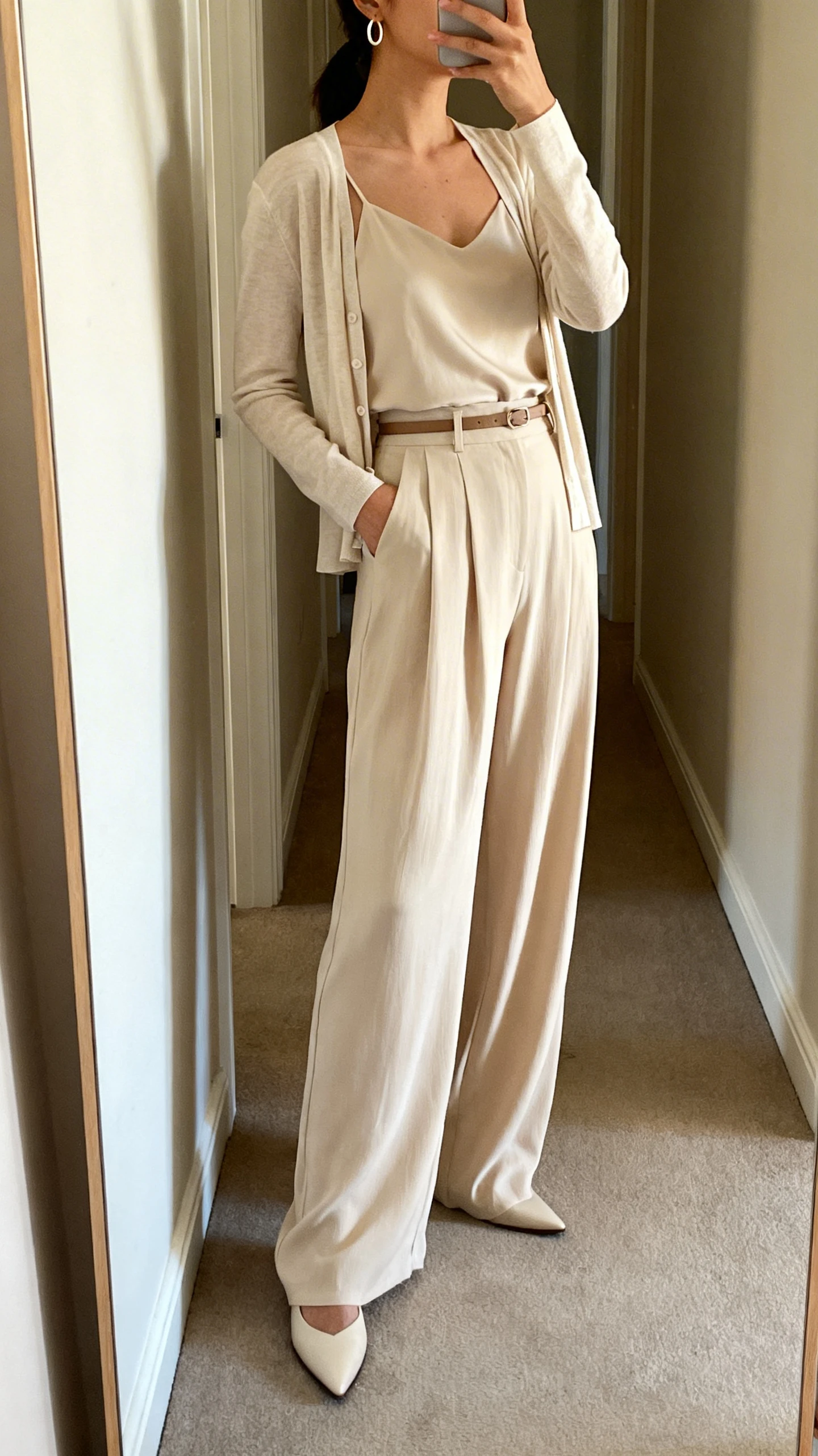 iPhone side-angle mirror selfie of a woman wearing a lightweight cardigan set (matching shell and cardigan), high-rise wide-leg drapey pants, pointed-toe flats, a thin belt, and small hoop earrings, face not visible, cropped at neck, full-length mirror in hallway, soft afternoon light, iPhone photo quality.