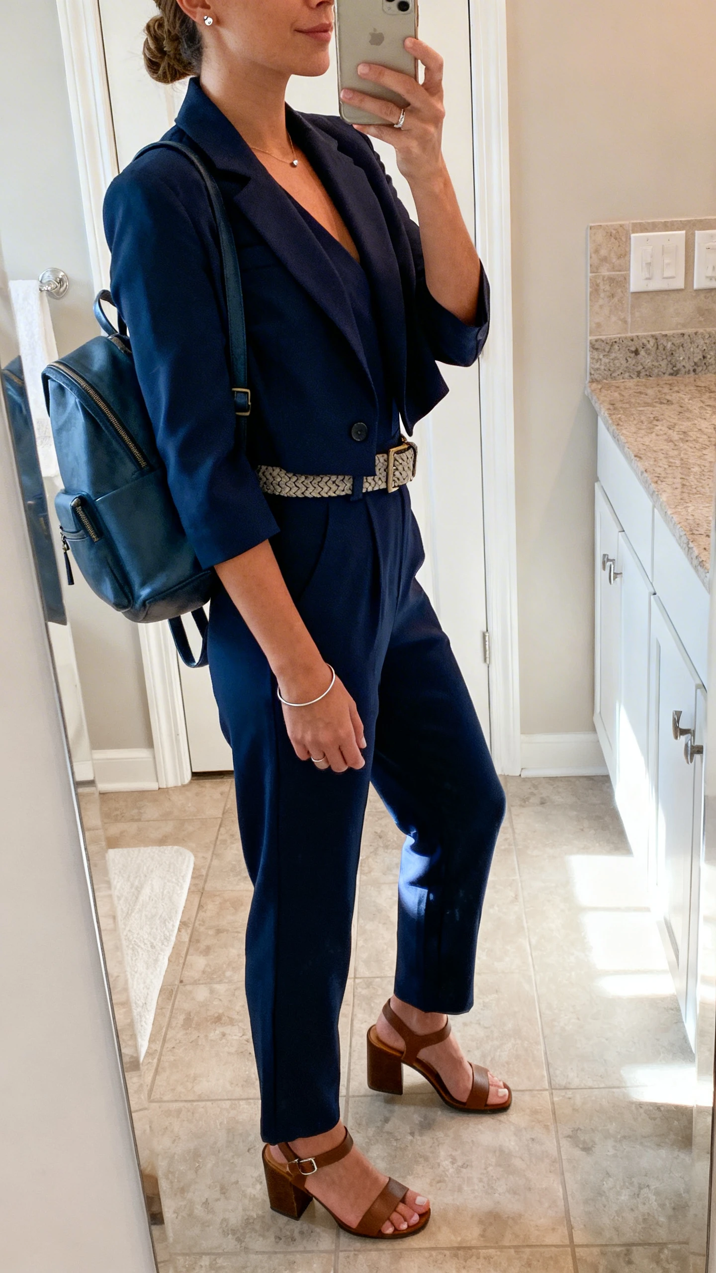 iPhone side-angle mirror selfie of a woman wearing a tailored navy jumpsuit with a lightweight cropped blazer, comfort block-heel sandals, a textured belt, subtle stud earrings, a thin bangle, and a medium backpack tote, face not visible, bathroom mirror, indoor morning light, iPhone photo quality.