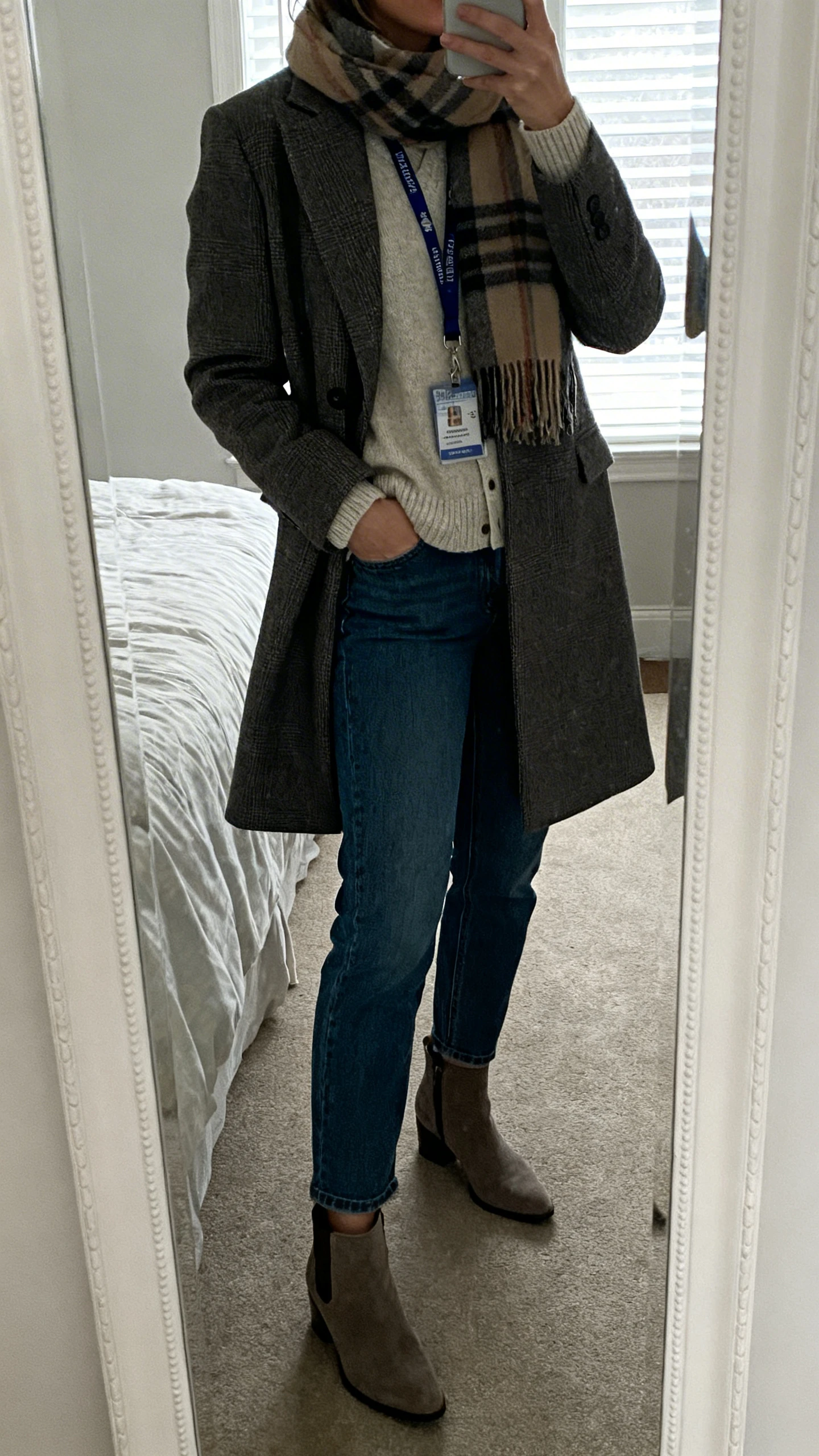 iPhone side-angle mirror selfie of a woman wearing a soft cardigan under a tailored coat, straight-leg jeans, ankle boots, scarf and teacher lanyard, face not visible, bedroom mirror, overcast window light for a cozy vibe, iPhone photo quality