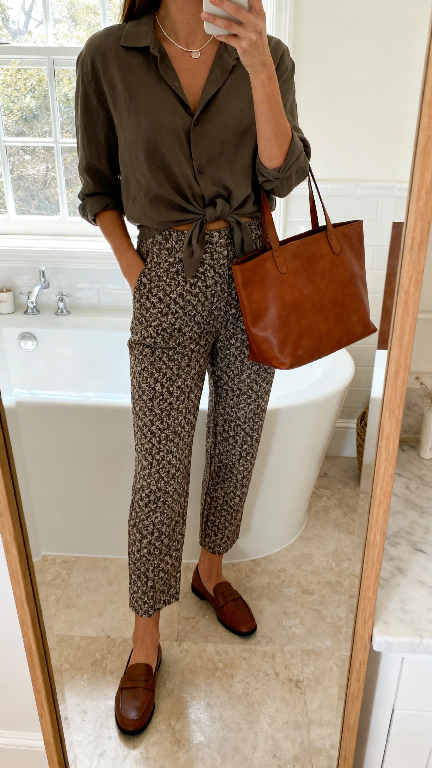 iPhone side-angle mirror selfie of a woman wearing patterned ankle-length trousers with a solid-color blouse half-tucked, loafers, minimalist necklace, leather tote, face not visible, bathroom mirror, natural window light, iPhone photo quality.