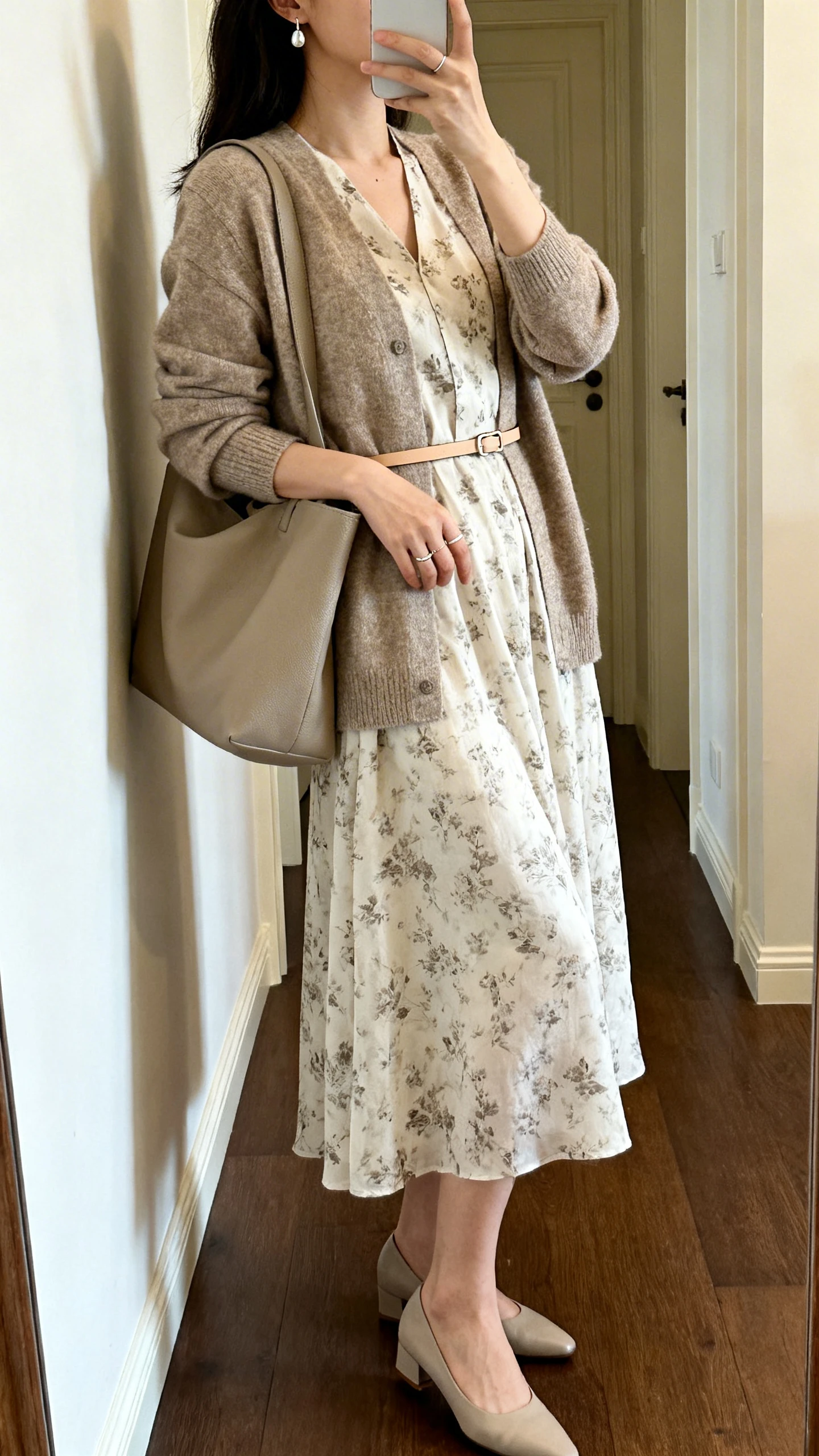 iPhone side-angle mirror selfie of a woman wearing a soft printed midi dress under a medium-weight cardigan cinched with a thin belt, comfort heels, subtle drop earrings, a minimal ring, and carrying a roomy tote, face not visible, cropped at neck, entryway mirror, natural indoor light, iPhone photo quality.