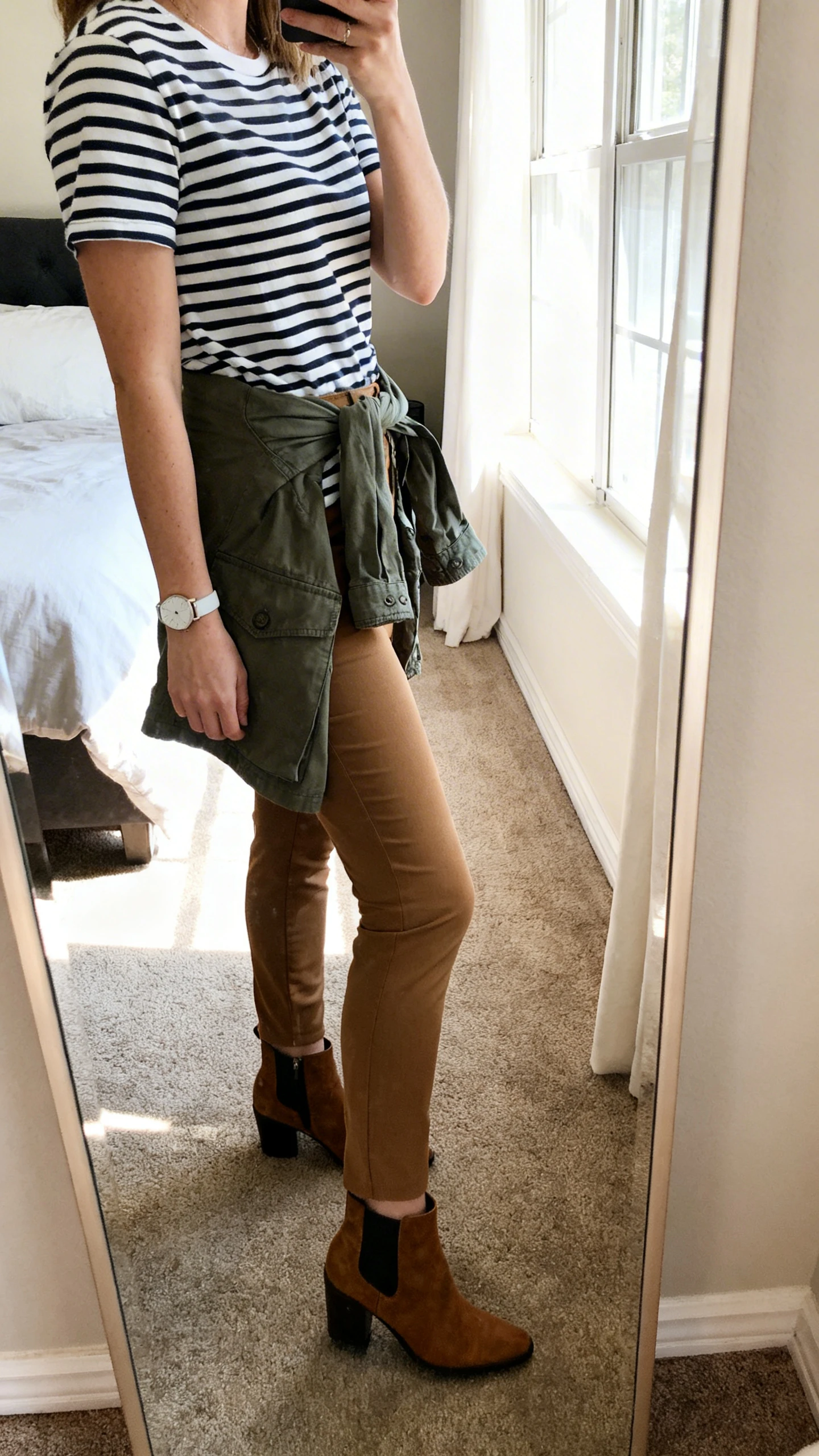 iPhone side-angle mirror selfie of a woman wearing stretch chinos, a Breton striped tee, ankle boots, utility jacket tied around hips, and a simple watch, face not visible, bedroom full-length mirror, afternoon window light, iPhone photo quality.
