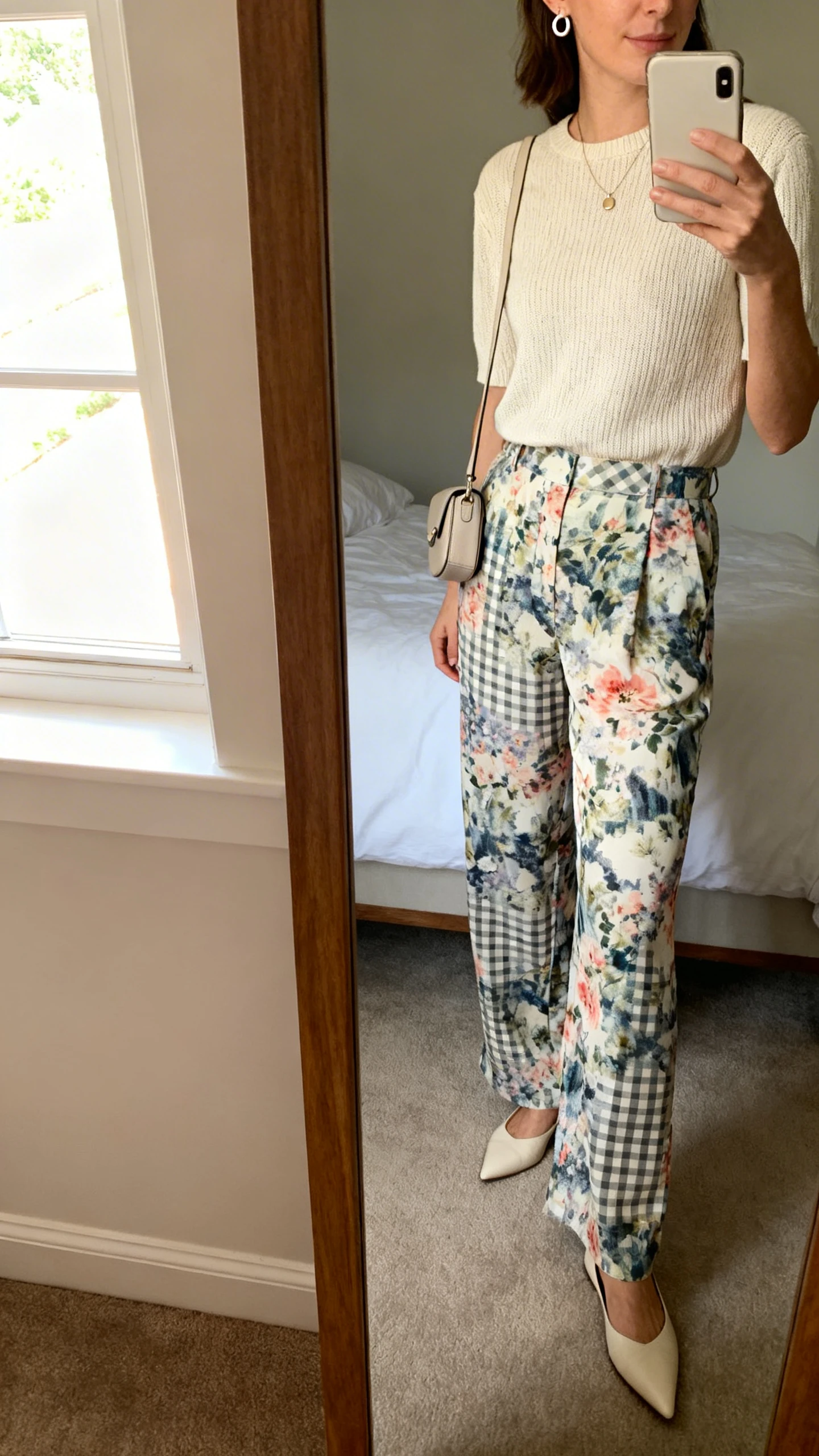 iPhone side-angle mirror selfie of a woman wearing lightweight printed trousers (abstract/floral/windowpane) with a solid fine-knit top, pointed flats, a subtle pendant, tiny huggie hoops, and a compact crossbody, face not visible, bedroom mirror with morning window light, iPhone photo quality.