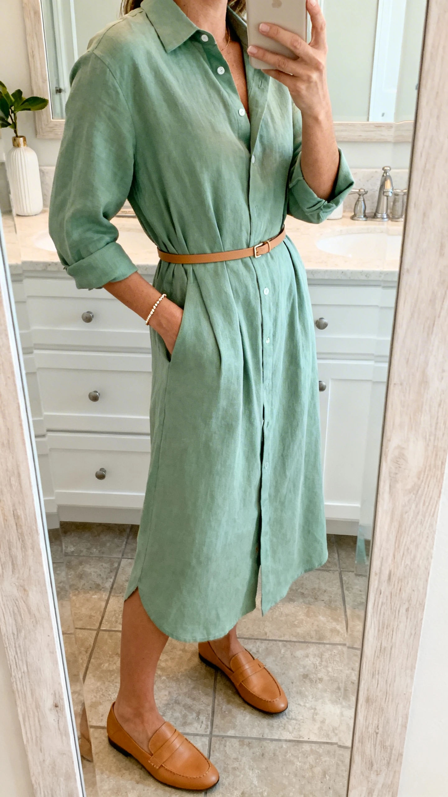 iPhone side-angle selfie of a woman wearing a soft shirt dress in calming sage green, cinched with a slim belt, paired with tan loafers and a delicate bracelet, face not visible, cropped at neck, bathroom mirror, gentle indoor lighting, iPhone photo quality.