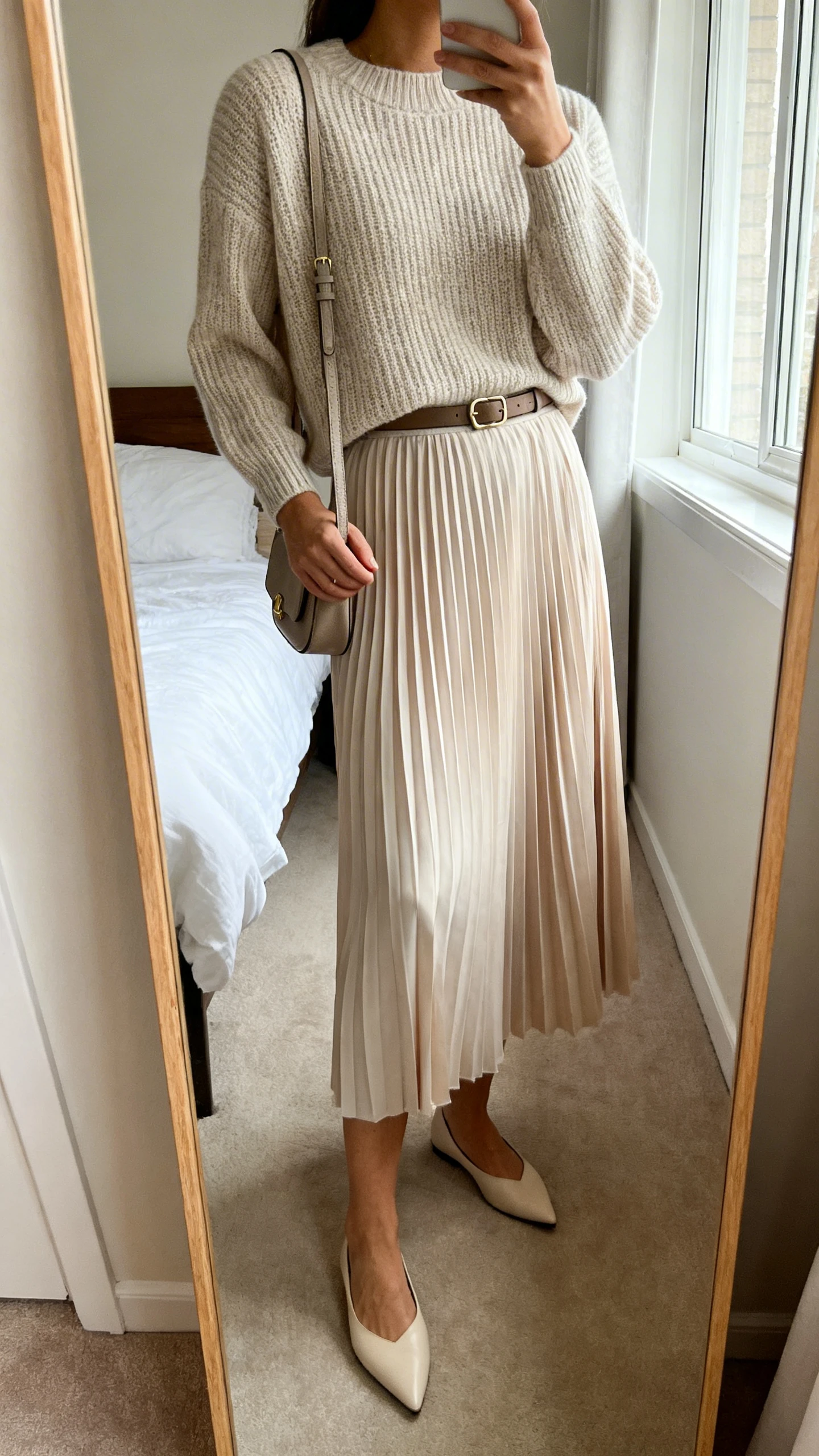 Side-angle iPhone mirror selfie of a woman wearing a fine-gauge knit sweater tucked into a flowy pleated midi skirt, pointed flats, slim belt, and small shoulder bag, face not visible, cropped at shoulders, bedroom mirror, natural window light, iPhone photo quality.
